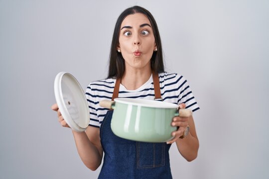 Young brunette woman wearing apron holding cooking pot making fish face with mouth and squinting eyes, crazy and comical.