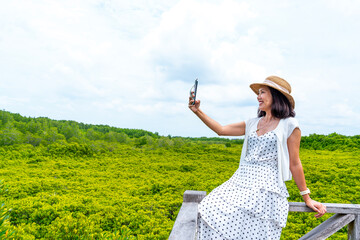 Tourist asian woman in white dress sitting on wooden bridge holding smartphone selfie in mangrove field in summer