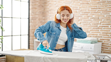 Young redhead woman listening to music ironing clothes dancing at laundry room