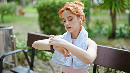 Fototapeta premium Young redhead woman using smartwatch and earphones sitting on bench at park