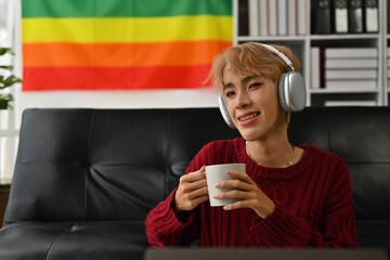 Portrait of attractive looking Asian gay teen boy smiling happily while relaxing listening to music on headphones and enjoying with hot drinks inside his home.