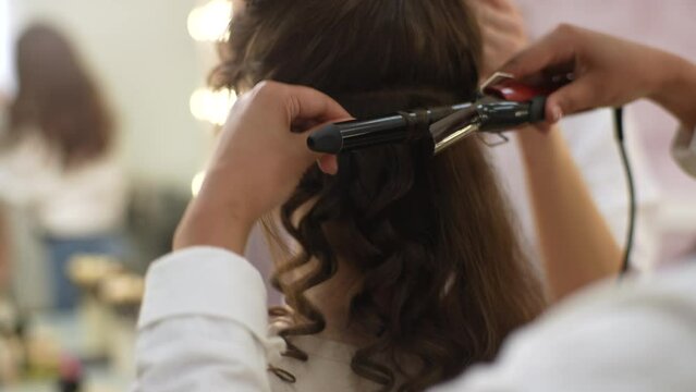 Closeup of process of curling long hair with hair tongs in modern beauty studio in front of mirror. Close up shot of female hairstylist curling hair using hair iron. Hairstylist doing stylish hairdo.