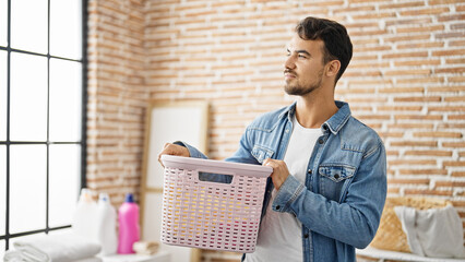 Young hispanic man holding basket with clothes at laundry room