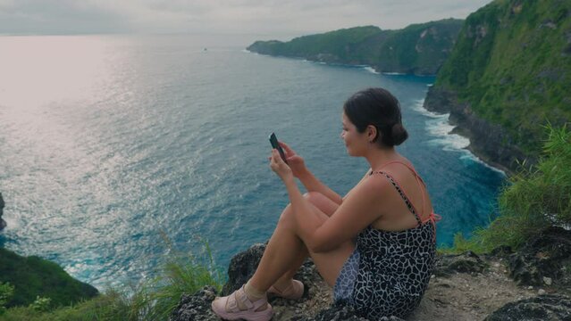 Asian girl relaxing on a rock above sea in summer with cell phone. Japanese woman holding hand phone sitting on stone at edge of cliff on ocean. Female hesitantly swipes his finger across phone screen