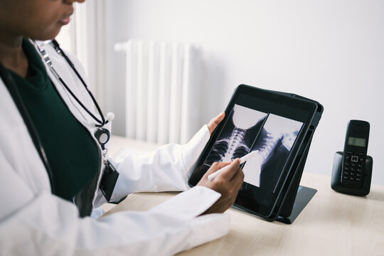 Crop black doctor sitting at table and checking x ray on tablet