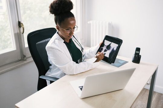 From above of black doctor sitting at table and showing x ray images on tablet