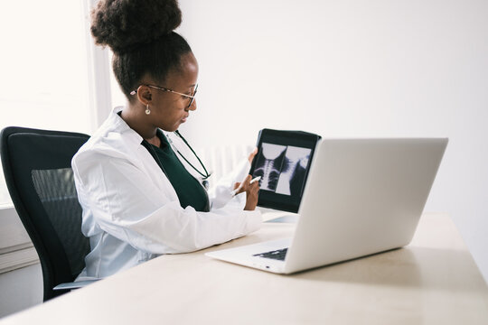 Side view of black doctor sitting at table and showing x ray images on tablet