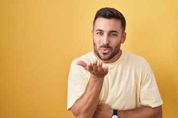 Handsome hispanic man standing over yellow background looking at the camera blowing a kiss with hand on air being lovely and sexy. love expression.