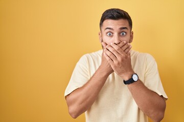 Handsome hispanic man standing over yellow background shocked covering mouth with hands for...