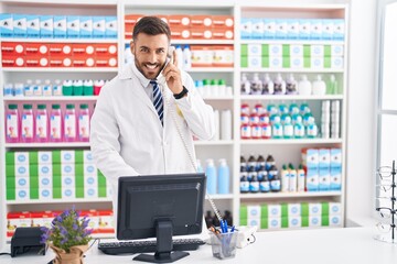 Young hispanic man pharmacist talking on telephone using computer at pharmacy