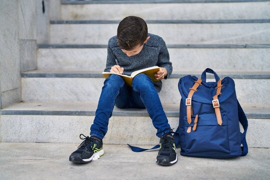 Blond child student writing on book sitting on stairs at school