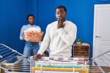 Young african american couple hanging clothes at clothesline covering mouth with hand, shocked and afraid for mistake. surprised expression
