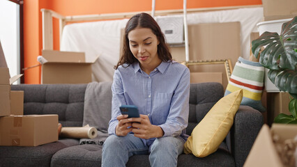 Obraz premium Young beautiful hispanic woman using smartphone sitting on the sofa at new home