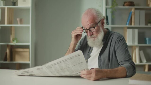 A Serious, Smart Old Man Sits at a Table and Reads a Newspaper.An Elderly Man with a Newspaper Alone at Home, Sees the News and Current Events in the Newspaper.Daily Newspaper Concept