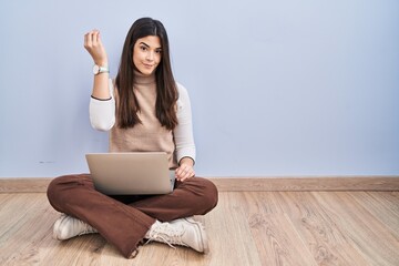 Young brunette woman working using computer laptop sitting on the floor doing italian gesture with hand and fingers confident expression