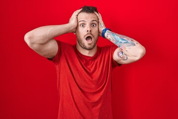 Young hispanic man standing over red background crazy and scared with hands on head, afraid and...