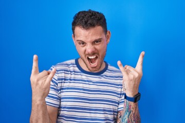 Young hispanic man standing over blue background shouting with crazy expression doing rock symbol with hands up. music star. heavy music concept.