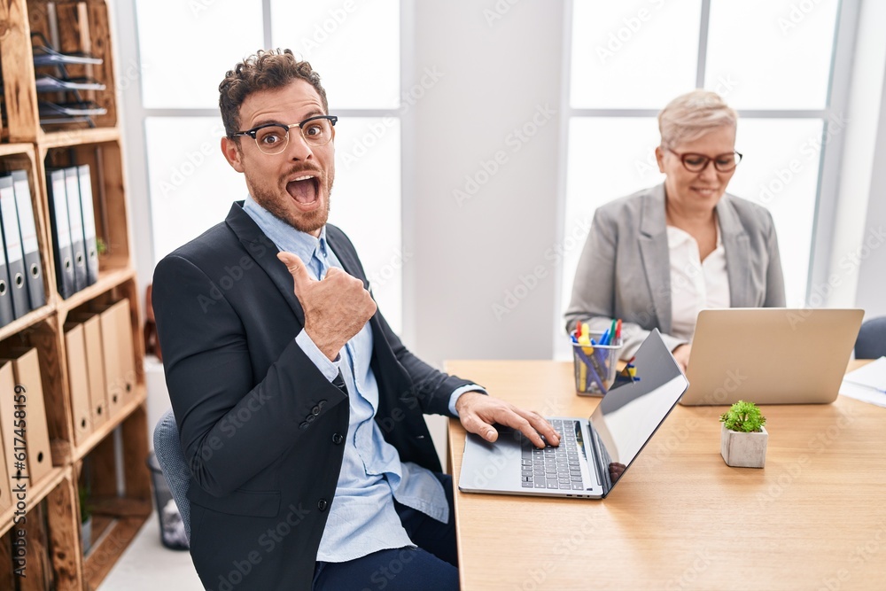 Wall mural Hispanic young man working at the office pointing thumb up to the side smiling happy with open mouth