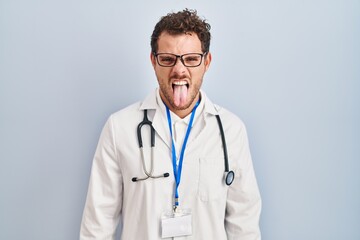 Young hispanic man wearing doctor uniform and stethoscope sticking tongue out happy with funny expression. emotion concept.