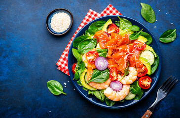 Fresh salad for keto diet with salmon, avocado, spinach, cucumber, shrimps, tomato, cashew, sesame seeds. Low-carbohydrate lunch rich in healthy fats. Blue stone table background, top view