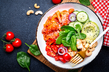 Keto diet salad with salmon, shrimp, avocado, spinach, cucumber, tomato, cashew, sesame. Low-carbohydrate lunch rich in healthy fats. Black table background, top view
