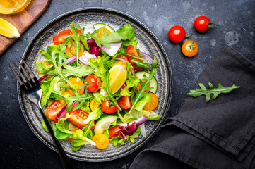 Summer vegan vegetable salad with colorful tomatoes, fresh cucumber, red onion, lettuce and arugula. Black table background, top view