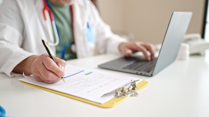 Middle age grey-haired man doctor using laptop writing medical report at clinic
