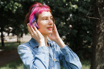 A girl with pink hair with headphones listens to music in the park in summer