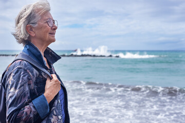 Portrait of senior gray haired woman in denim jacket and backpack standing in front to the sea...