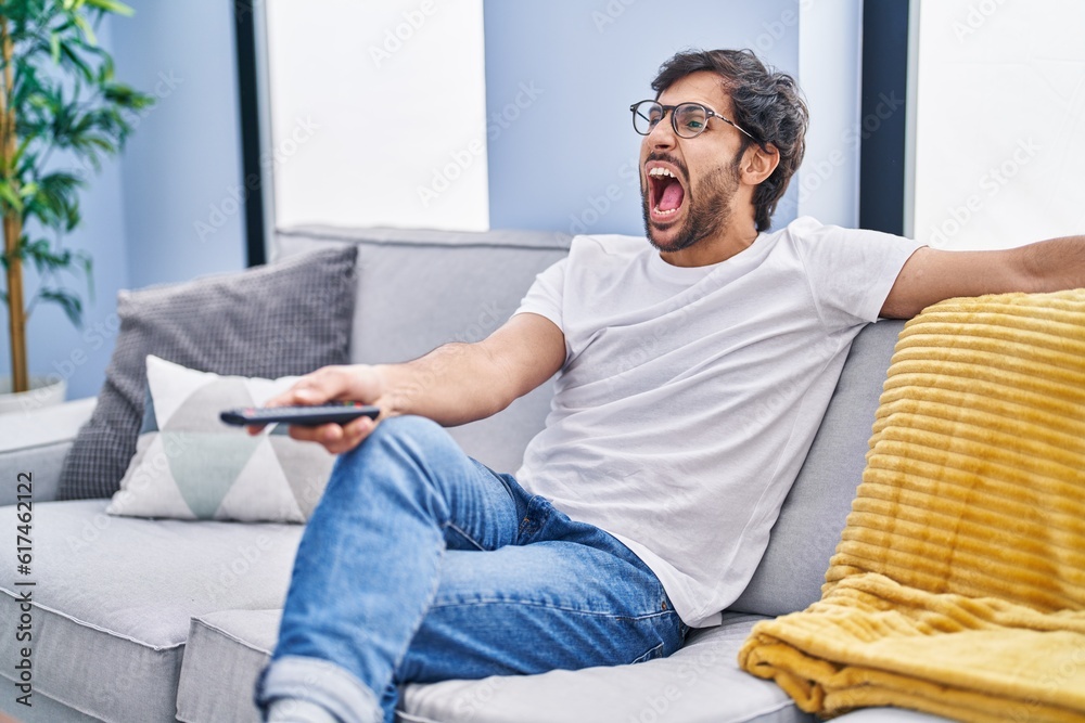 Sticker Handsome latin man holding television remote control angry and mad screaming frustrated and furious, shouting with anger. rage and aggressive concept.