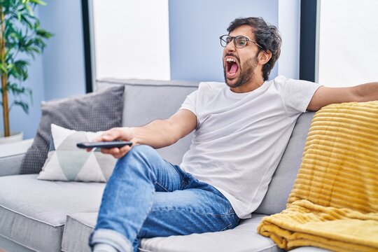 Handsome Latin Man Holding Television Remote Control Angry And Mad Screaming Frustrated And Furious, Shouting With Anger. Rage And Aggressive Concept.