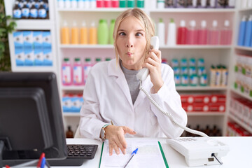 Young caucasian woman working at pharmacy drugstore speaking on the telephone making fish face with lips, crazy and comical gesture. funny expression.