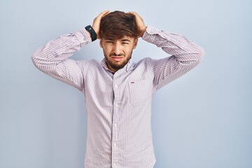 Arab man with beard standing over blue background suffering from headache desperate and stressed because pain and migraine. hands on head.