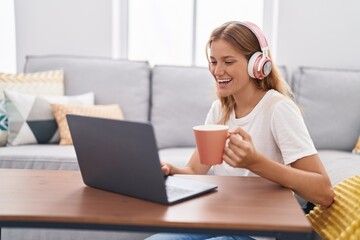 Young blonde girl watching video on laptop drinking coffee at home