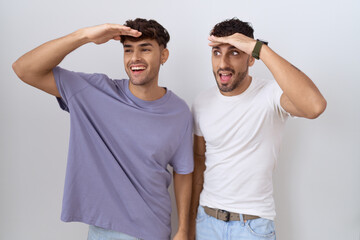 Homosexual gay couple standing over white background very happy and smiling looking far away with hand over head. searching concept.