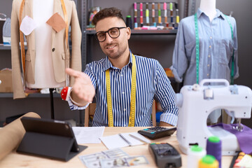 Hispanic man with beard dressmaker designer working at atelier smiling cheerful offering palm hand giving assistance and acceptance.