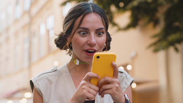 Oh my God Wow. Excited happy joyful winner pretty young woman use smartphone typing browsing celebrating win good message news. Girl with mobile phone walking in urban summer city street outdoors