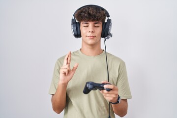 Hispanic teenager playing video game holding controller gesturing finger crossed smiling with hope...