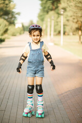 Cute little girl in protective equipment and rollers stands on walkway in park outdoor. Summer activity for children.