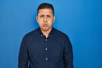 Hispanic young man standing over blue background puffing cheeks with funny face. mouth inflated...