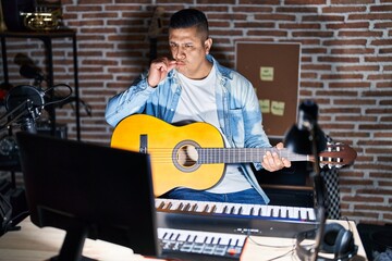 Hispanic young man playing classic guitar at music studio mouth and lips shut as zip with fingers....