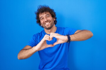 Hispanic young man standing over blue background smiling in love doing heart symbol shape with...