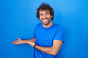 Hispanic young man standing over blue background inviting to enter smiling natural with open hand