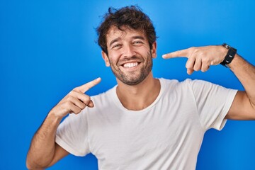 Hispanic young man standing over blue background smiling cheerful showing and pointing with fingers teeth and mouth. dental health concept.