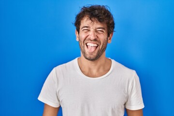 Fototapeta premium Hispanic young man standing over blue background sticking tongue out happy with funny expression. emotion concept.
