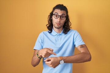 Young hispanic man standing over yellow background in hurry pointing to watch time, impatience,...