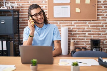 Young hispanic man business worker using laptop talking on smartphone at office