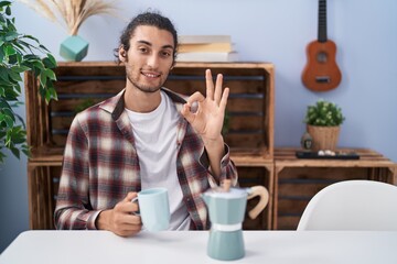 Young hispanic man drinking coffee from french coffee maker doing ok sign with fingers, smiling...