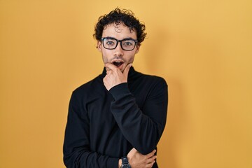 Hispanic man standing over yellow background looking fascinated with disbelief, surprise and amazed expression with hands on chin