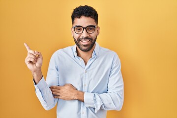 Hispanic man with beard standing over yellow background with a big smile on face, pointing with hand and finger to the side looking at the camera.